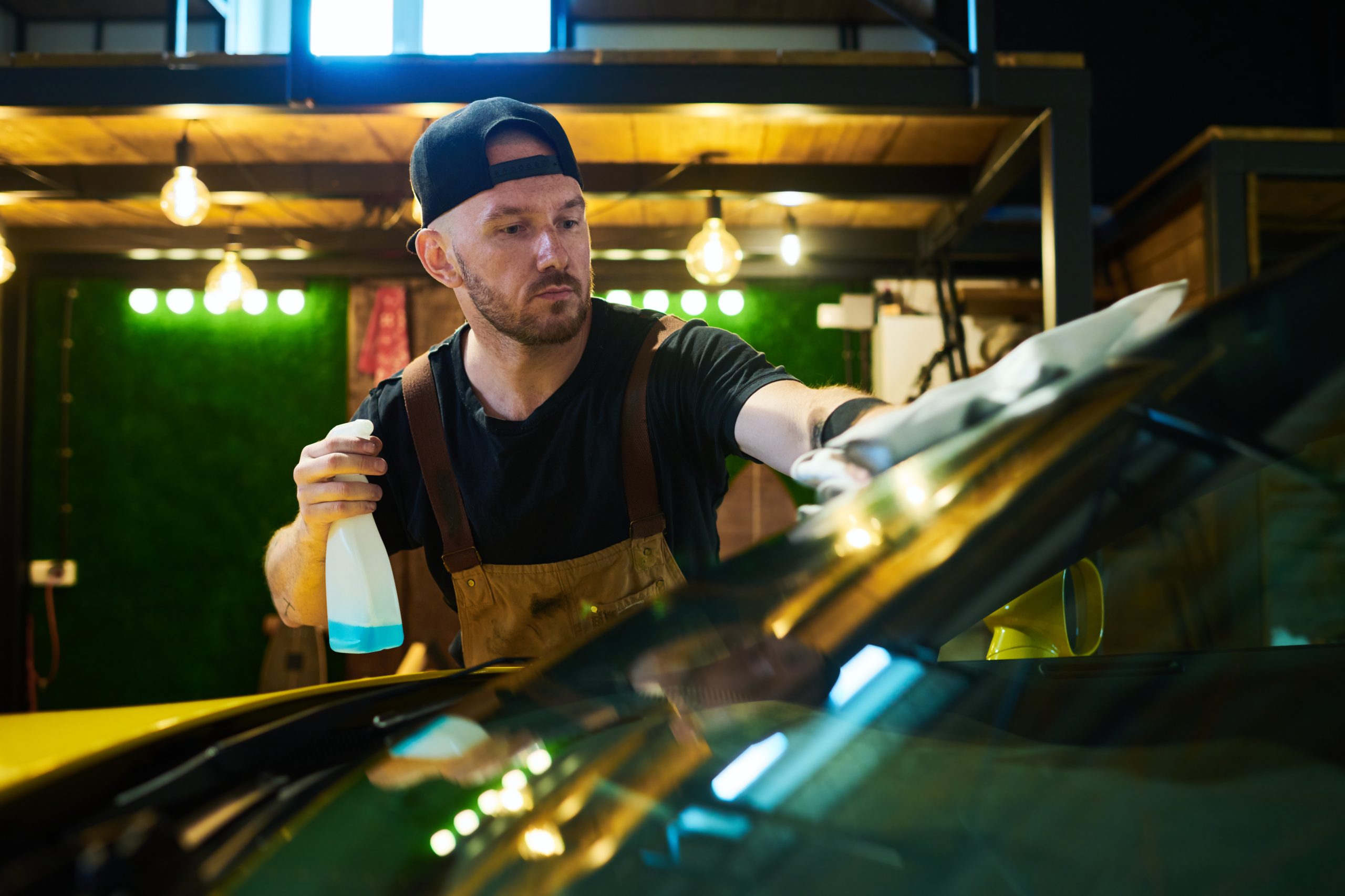 Young male worker of maintenance service holding plastic bottle with sprayer and wiping windscreen with duster while cleaning it